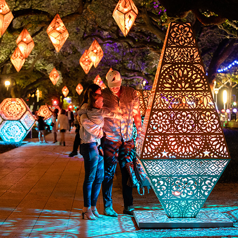 Family interacting with large geometric sculpture lit from within amid hanging geometric lanterns in a garden setting